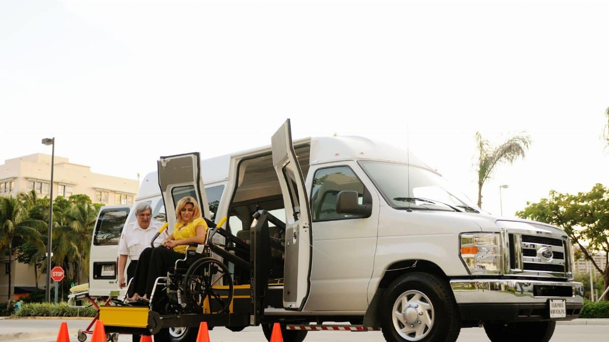 A woman in a wheelchair uses a lift to board an accessibility van assisted by a driver.