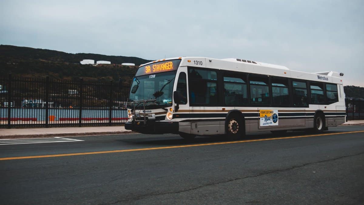 Big gray bus driving on asphalt highway along metal fence near water against cloudless sky with mountains in distance outside