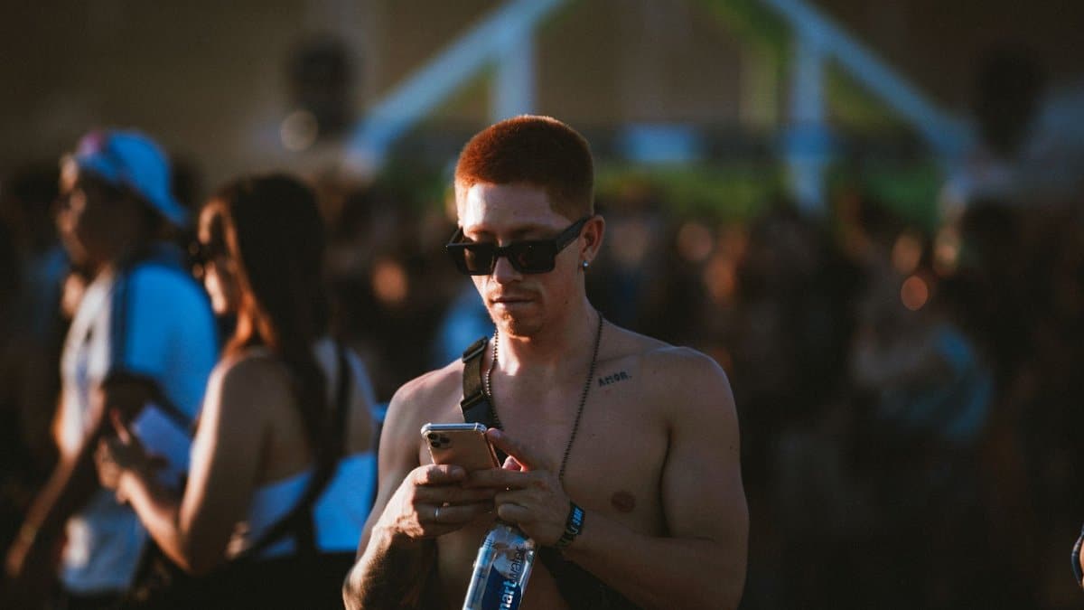 Shirtless man wearing sunglasses at a lively outdoor music festival, checking phone.