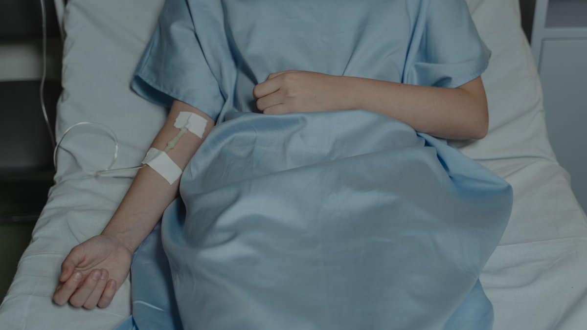 A patient lies in bed wearing a hospital gown with an IV drip attached, evoking a sense of illness.