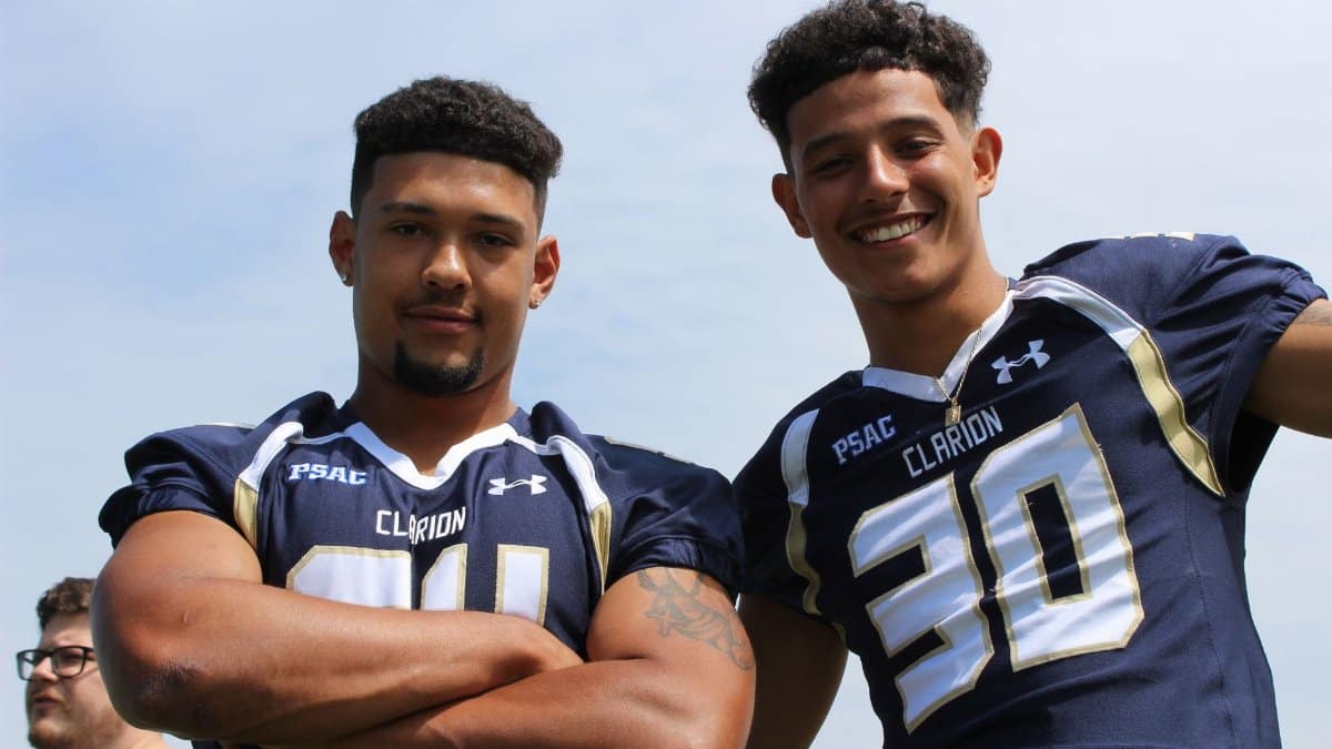 Two young American football players in uniform smiling confidently outdoors.