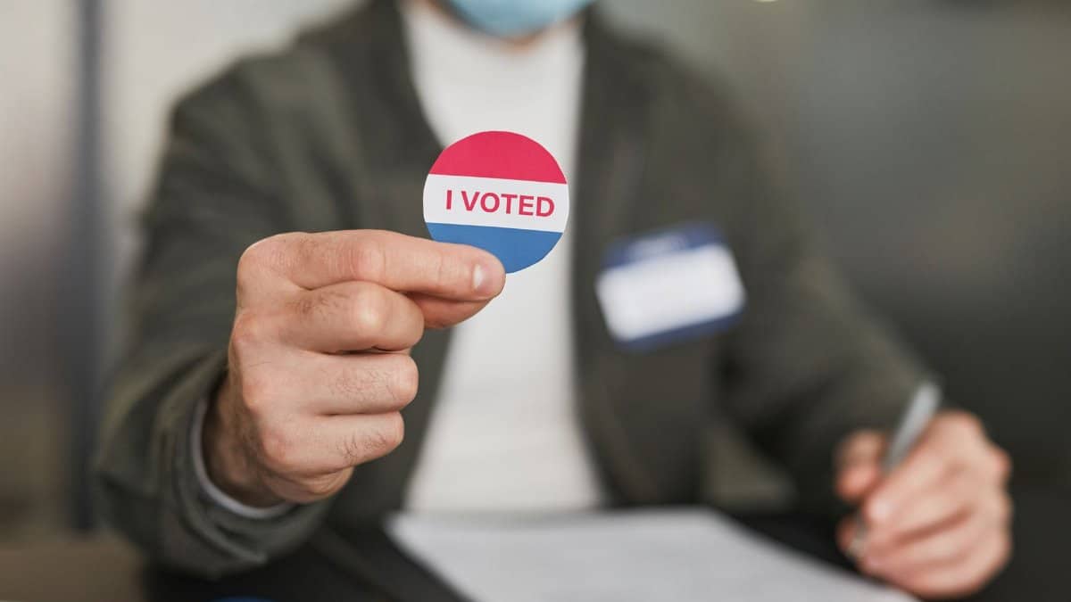 A person in a mask holding an 'I Voted' sticker, emphasizing civic engagement.