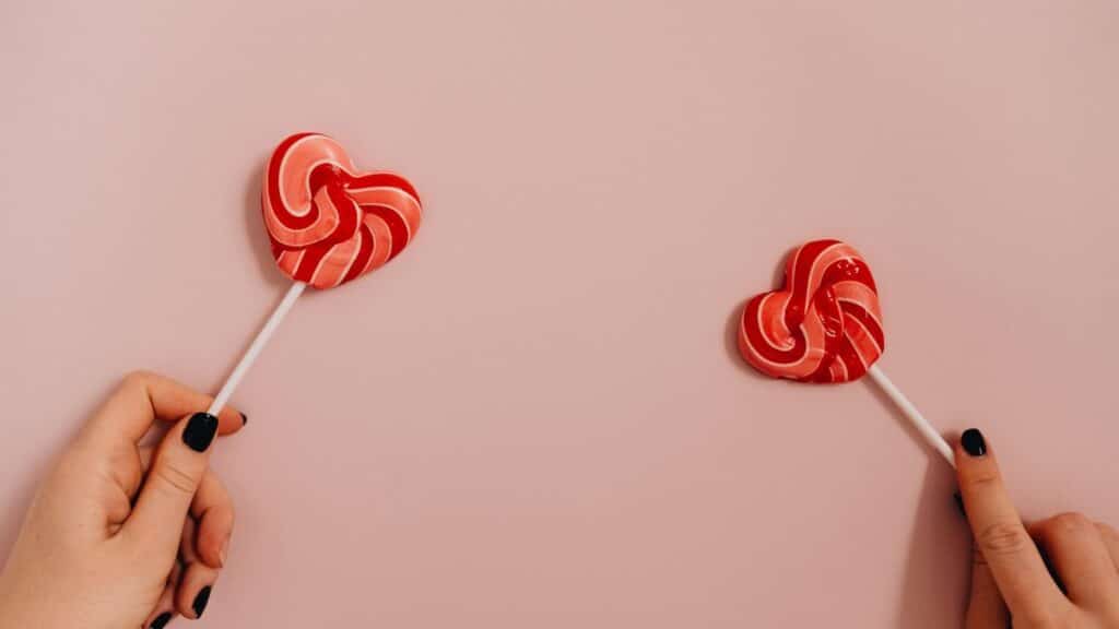 Two hands holding heart-shaped lollipops against a pastel pink background.