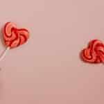 Two hands holding heart-shaped lollipops against a pastel pink background.