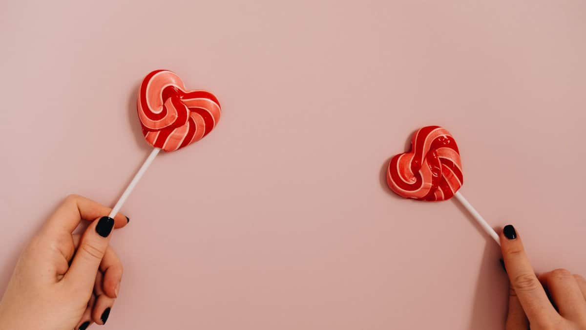 Two hands holding heart-shaped lollipops against a pastel pink background.
