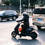 A person on a scooter navigates city traffic alongside cars in a busy urban street.