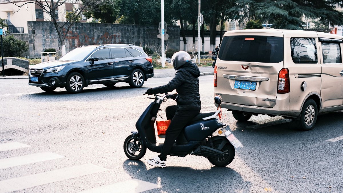 A person on a scooter navigates city traffic alongside cars in a busy urban street.