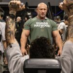 A gym trainer oversees a client's workout on a machine as others observe the session.