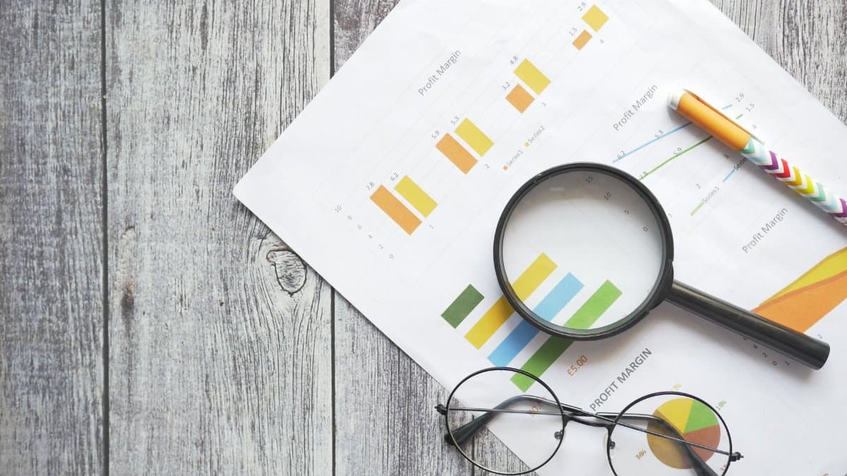 Overhead view of financial charts, a magnifying glass, ballpen, and eyeglasses on a wooden surface.