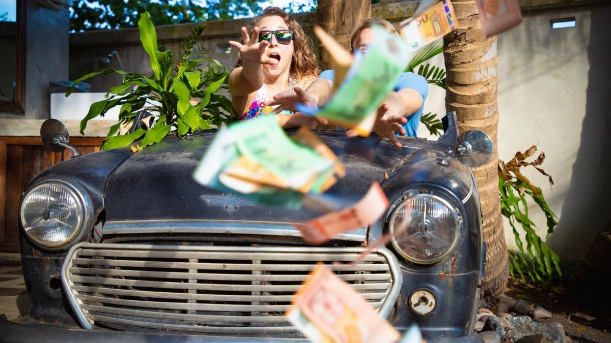 Two people reach for falling money in front of a vintage car in an outdoor setting.