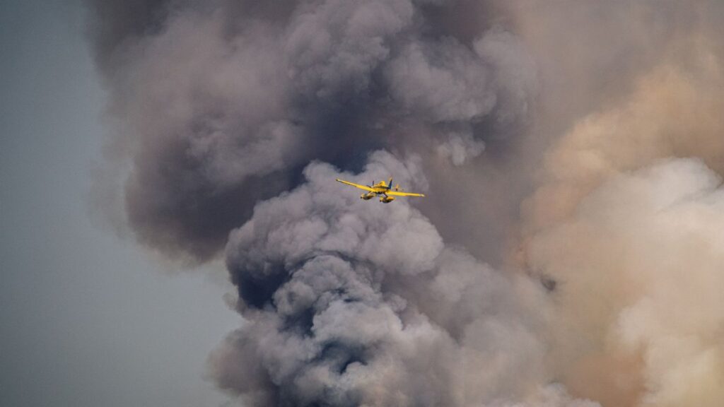 Dramatic scene of a yellow airplane flying through dense smoke, possibly from a wildfire.