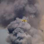 Dramatic scene of a yellow airplane flying through dense smoke, possibly from a wildfire.