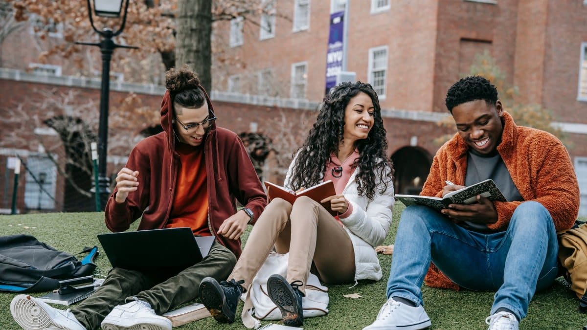 Full body of happy diverse students with notebooks and laptop sitting on grassy lawn on campus of university while studying together