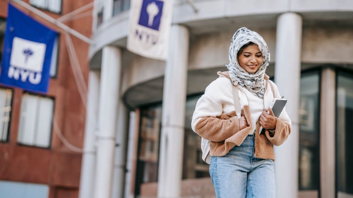 Low angle of cheerful young Arab female student in stylish warm clothes and hijab standing on city street near university building with copybooks and backpack and smiling