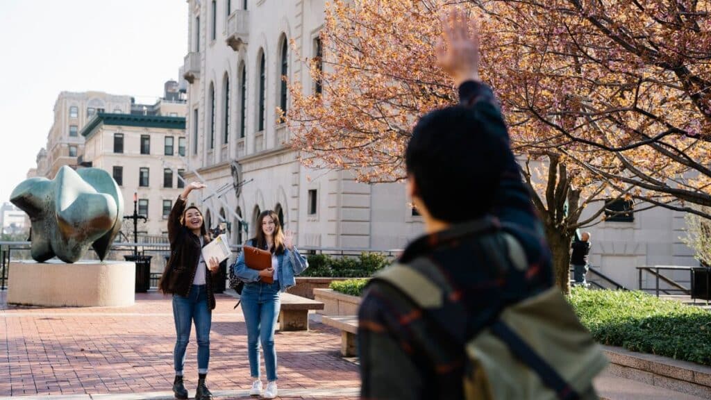 Two university students greet a friend in a vibrant autumn plaza lined with cherry blossom trees.