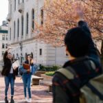 Two university students greet a friend in a vibrant autumn plaza lined with cherry blossom trees.