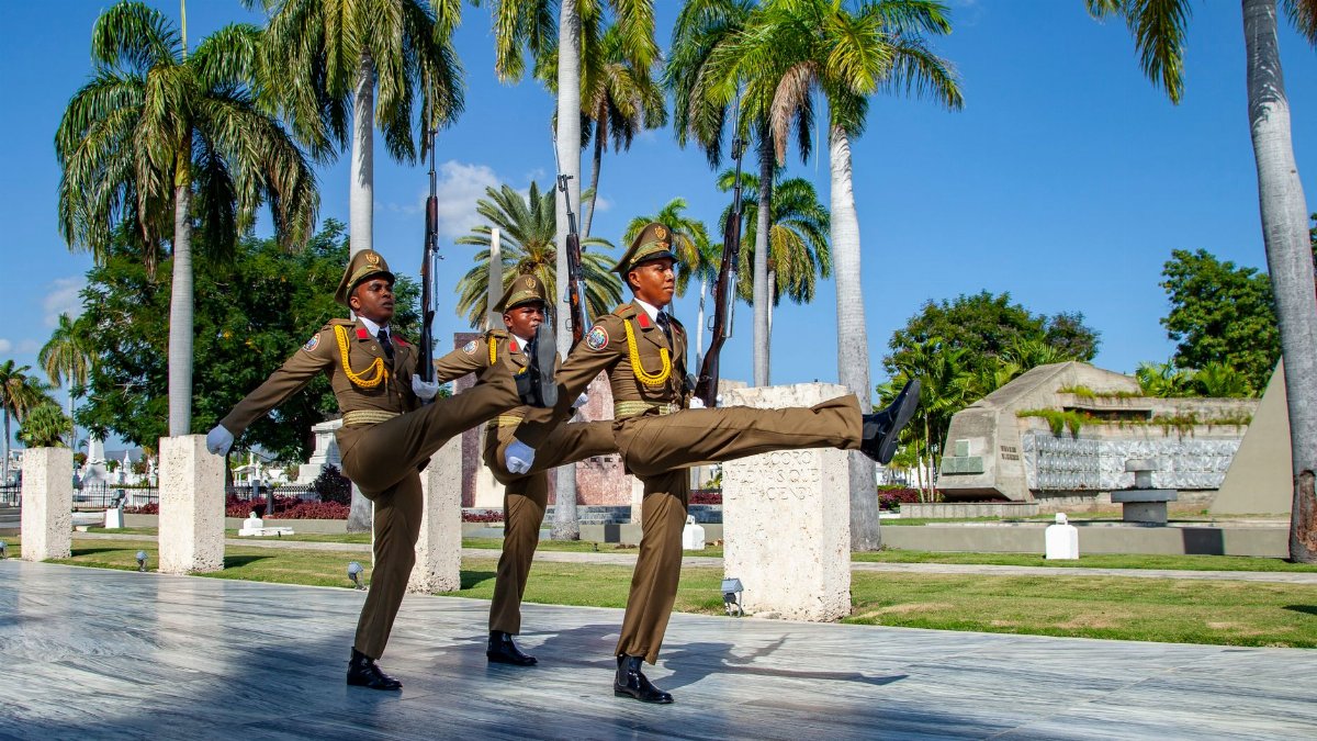 Military personnel performing a ceremonial march in Santiago de Cuba, surrounded by palm trees.