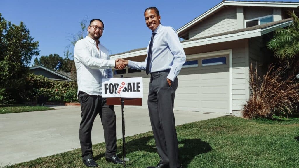 Two men shake hands in front of a new home with a sold sign, symbolizing a successful real estate deal.