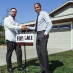 Two men shake hands in front of a new home with a sold sign, symbolizing a successful real estate deal.