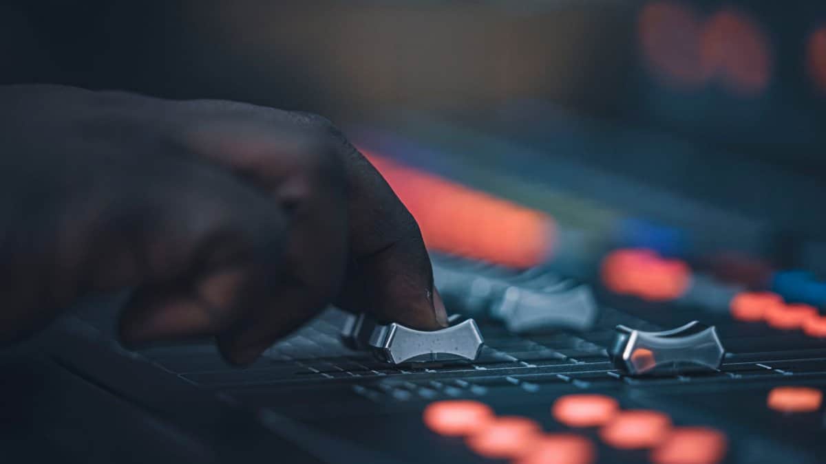 A hand adjusting sliders on a professional audio mixing console in a dimly lit studio setup.