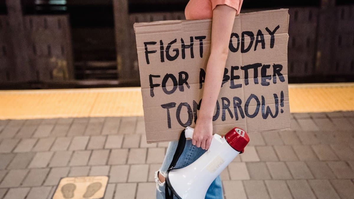 A woman stands in a subway holding a cardboard protest sign and megaphone, advocating for change.