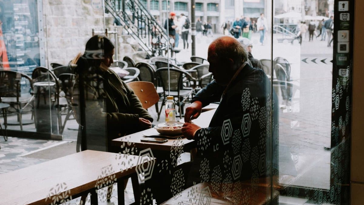 Silhouette of two people dining inside a cafe through a window with reflections of city life outside.
