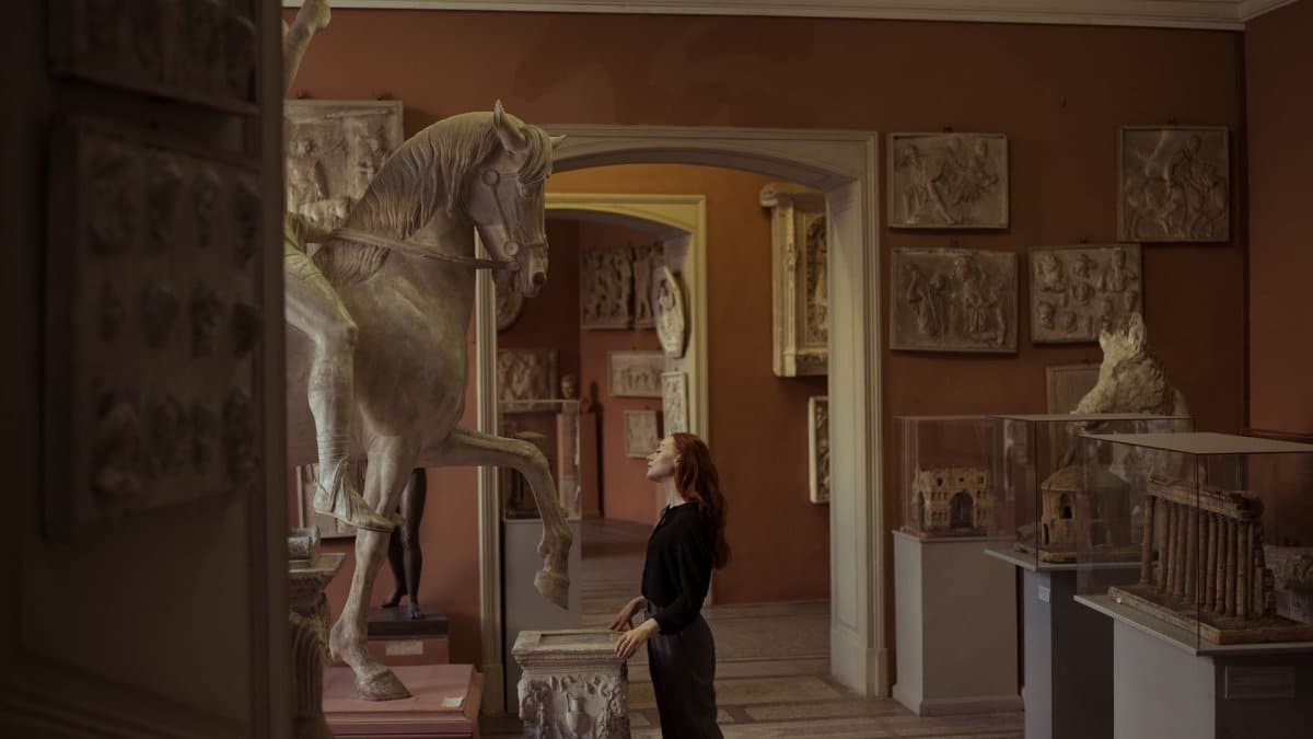 Woman admiring a horse sculpture in an art museum, surrounded by classical artworks.