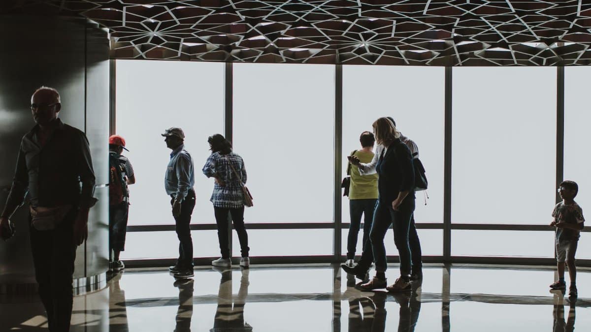 Silhouettes of tourists observing the Dubai skyline from an indoor observation deck.