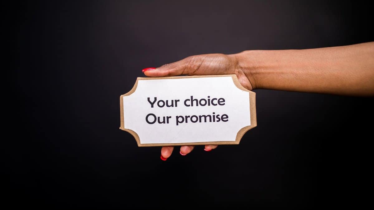 A hand holds a sign saying 'Your choice, Our promise' against a dark background.