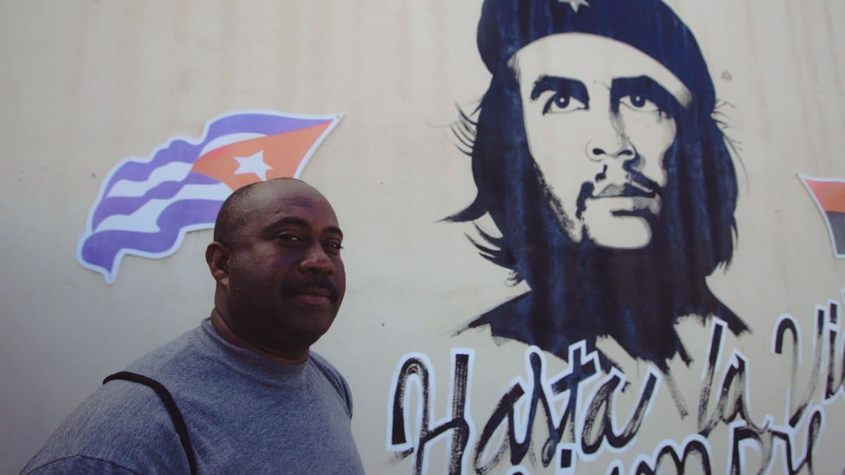African American man with Che Guevara mural and Cuban flag in the background, outdoor setting.