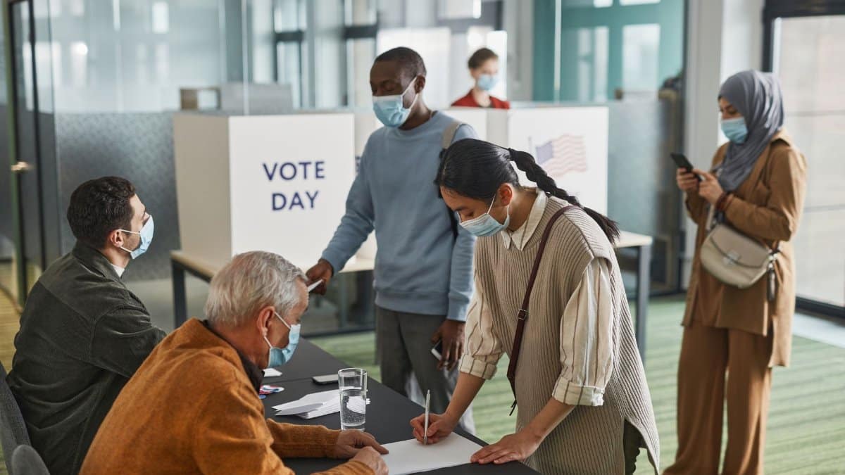 A diverse group of adults casting votes indoors on election day, maintaining safety protocols.