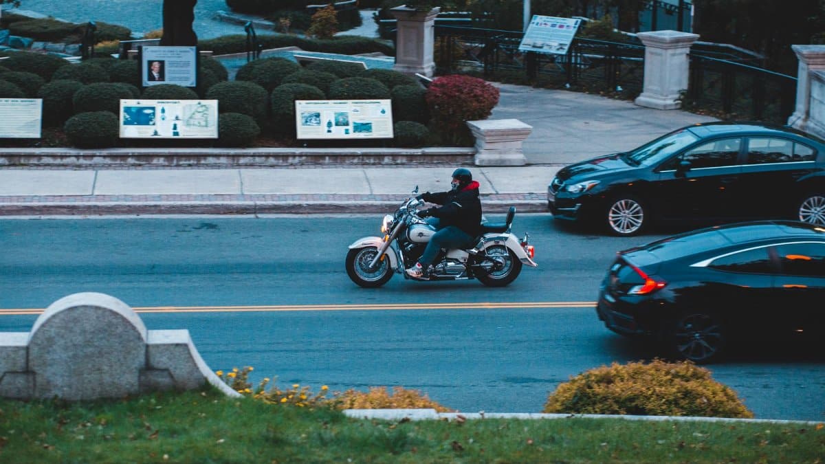 From above of anonymous male biker riding motorcycle on asphalt road with cars along grassy park in city street outside