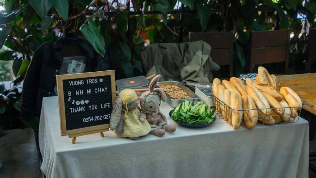 A welcoming cafe table set with Banh Mi ingredients and an inviting sign in a leafy atmosphere.