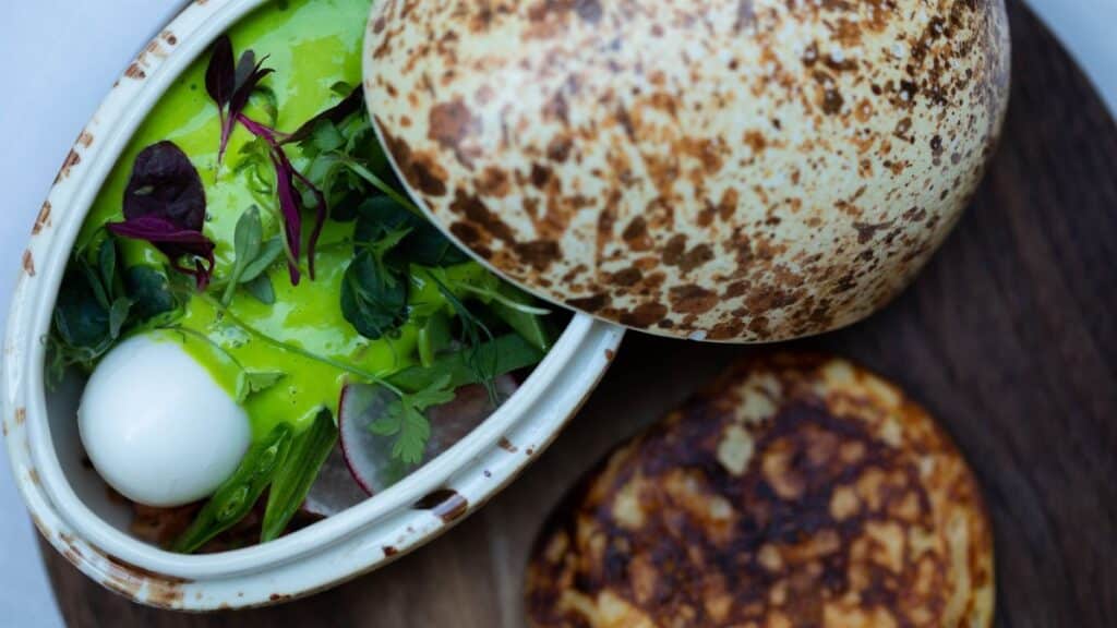 From above of appetizing vegetable and greenery and cheese salad with pesto sauce in pottery with open lid served with flatbread on wooden board