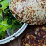 From above of appetizing vegetable and greenery and cheese salad with pesto sauce in pottery with open lid served with flatbread on wooden board