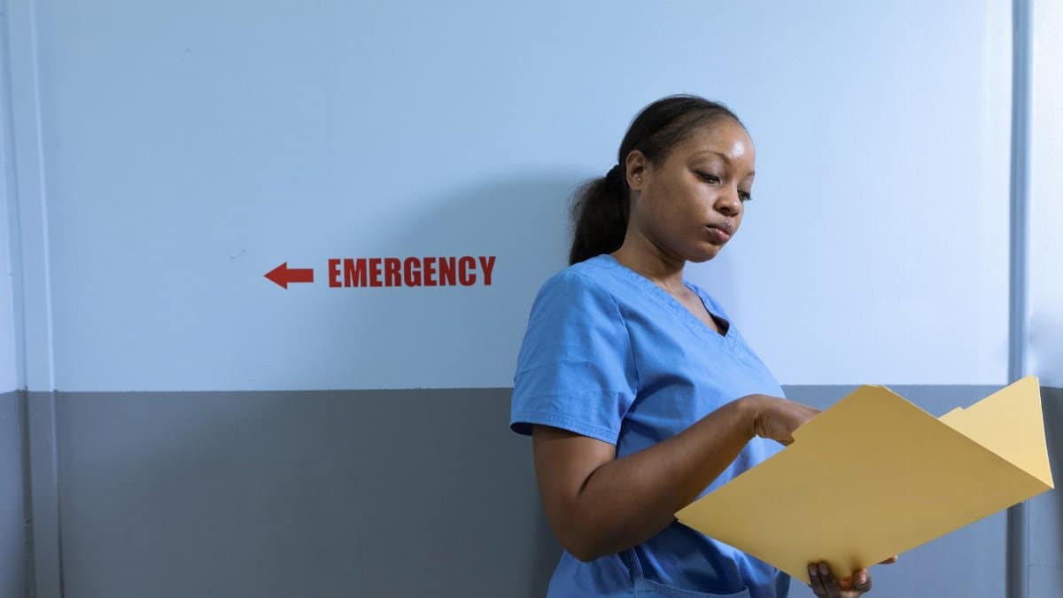 A nurse in blue scrubs reads a folder near an emergency room sign in a hospital.