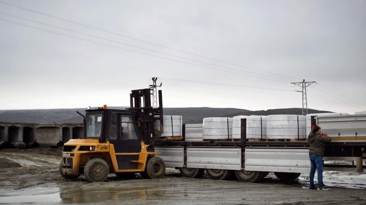 A forklift is loading cargo onto a truck in a muddy logistics area with a worker assisting.
