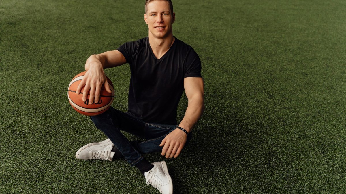 Muscular man sitting on grass holding basketball, wearing casual attire.