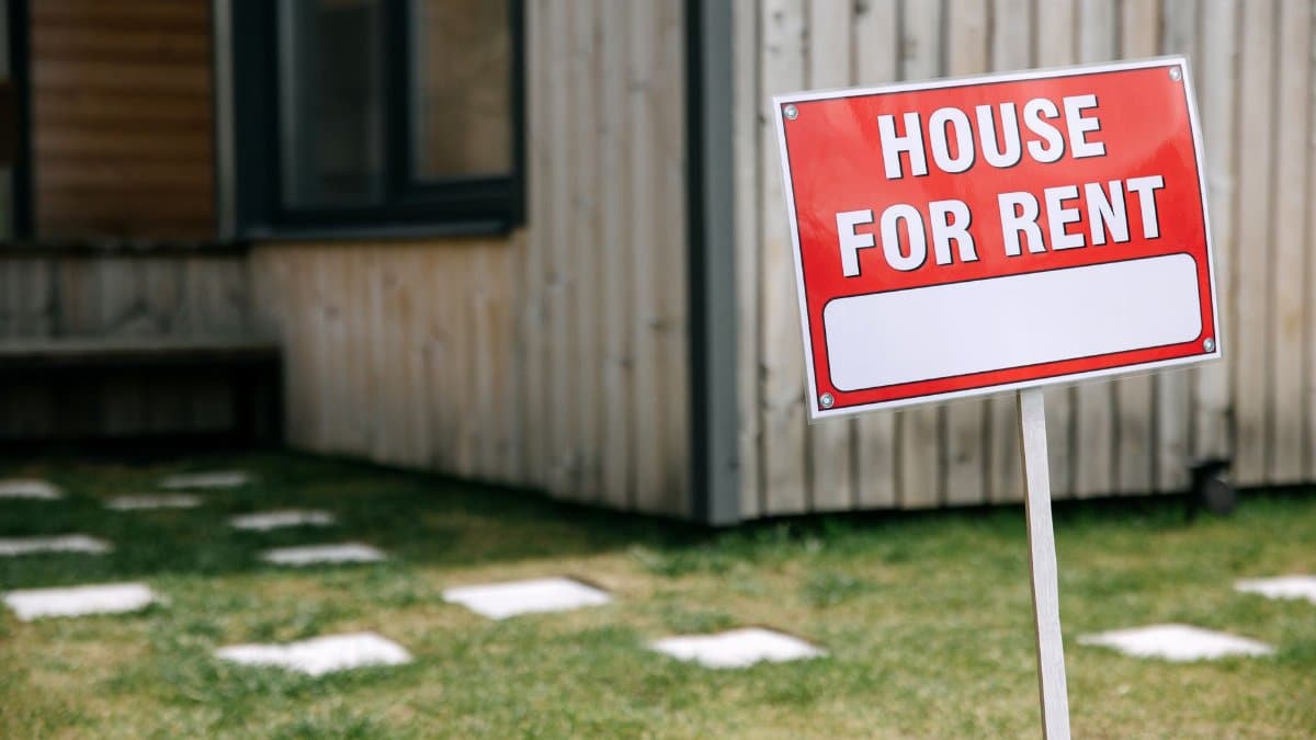 A modern wooden house with a prominent 'House for Rent' sign in the green yard.