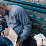 A woman compassionately offers food to a homeless man sleeping on a bench with a 'Hungry' sign.