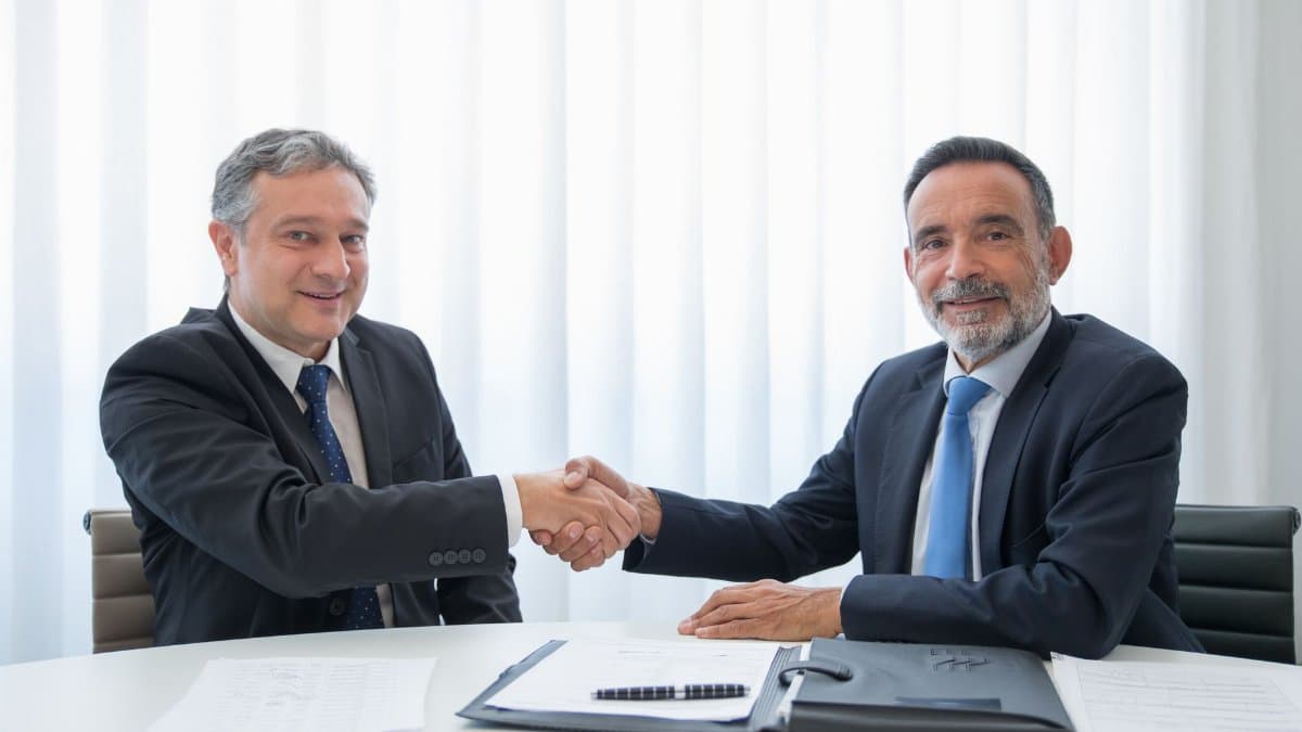 Two businessmen in suits shaking hands at an office table with documents.
