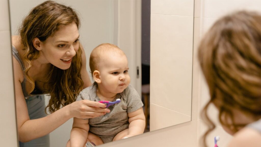 A mother helps her baby brush teeth while they share a joyful moment in front of a bathroom mirror.