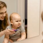 A mother helps her baby brush teeth while they share a joyful moment in front of a bathroom mirror.