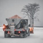 A snowplow truck spreading salt on a snowy road during a winter storm, with bare trees in the background.