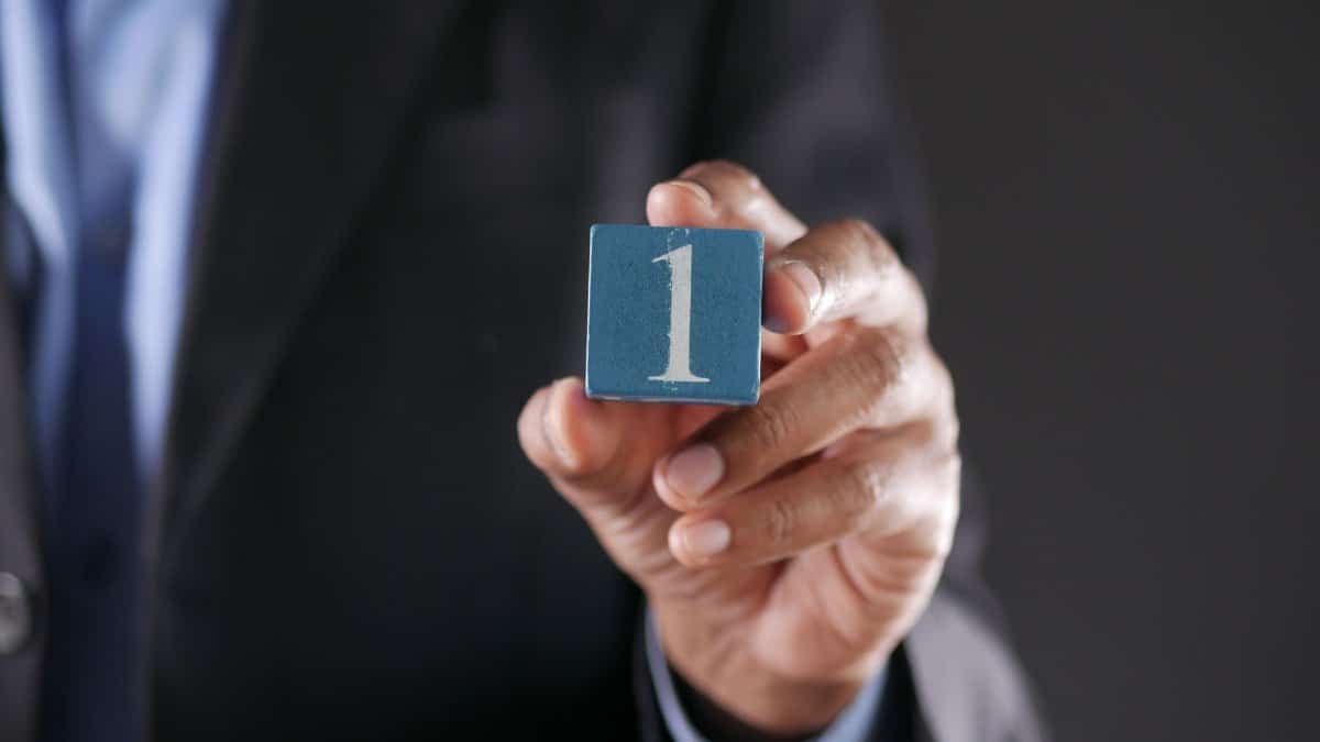 A hand in a suit holds a blue block with number one on a dark background.