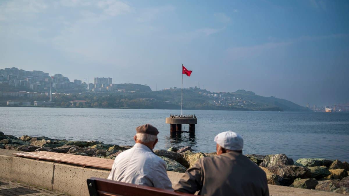 Two elderly men sit on a bench by the sea, viewing a distant flag over calm waters.