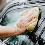 A hand scrubs a car window with a sponge, covered in soap suds, during a car wash.
