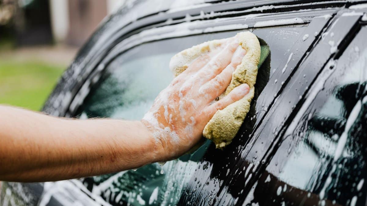 A hand scrubs a car window with a sponge, covered in soap suds, during a car wash.