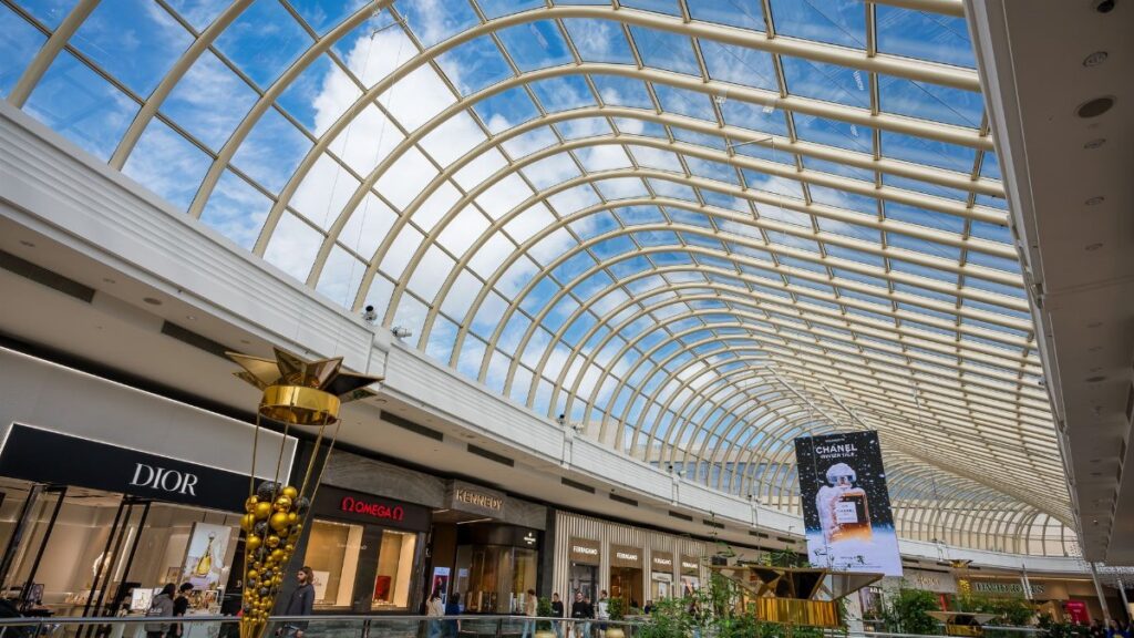 Inside a modern shopping mall with a stunning glass roof and luxury brand stores under a vibrant blue sky.