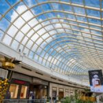 Inside a modern shopping mall with a stunning glass roof and luxury brand stores under a vibrant blue sky.